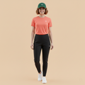 Woman wearing an orange shirt, black leggings, and a green cap on a beige background